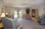 A cozy living room in a nursing home with several upholstered chairs arranged around a television set in a wooden entertainment center. The room has carpeted floors, framed pictures on the walls, a floor lamp, and a large potted plant near glass doors that lead outside.