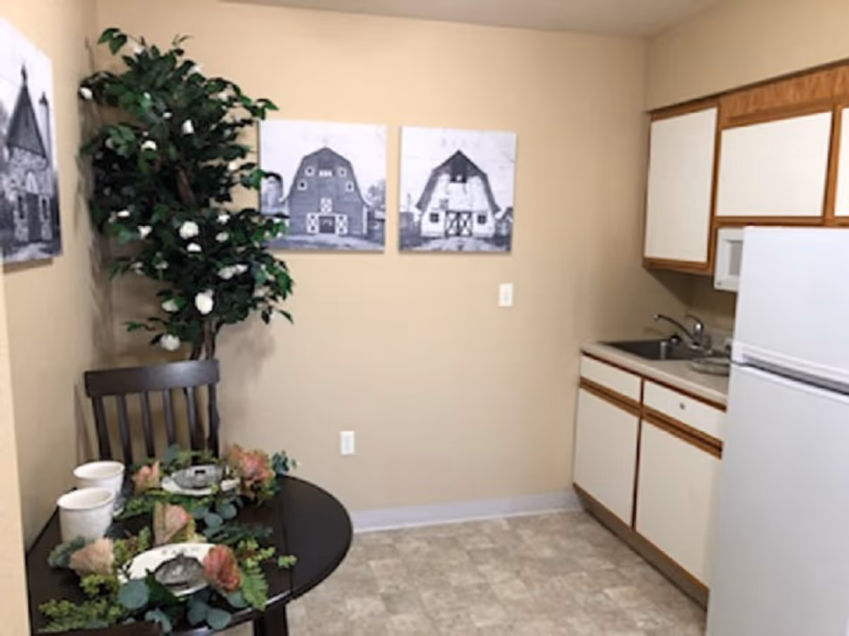 Small kitchen area with white cabinets and a white refrigerator on the right side. A round black table with two place settings, cups, and floral decorations is on the left side next to a wooden chair. There is a tall green plant with white flowers in the corner and three black and white barn-themed pictures on the beige wall.