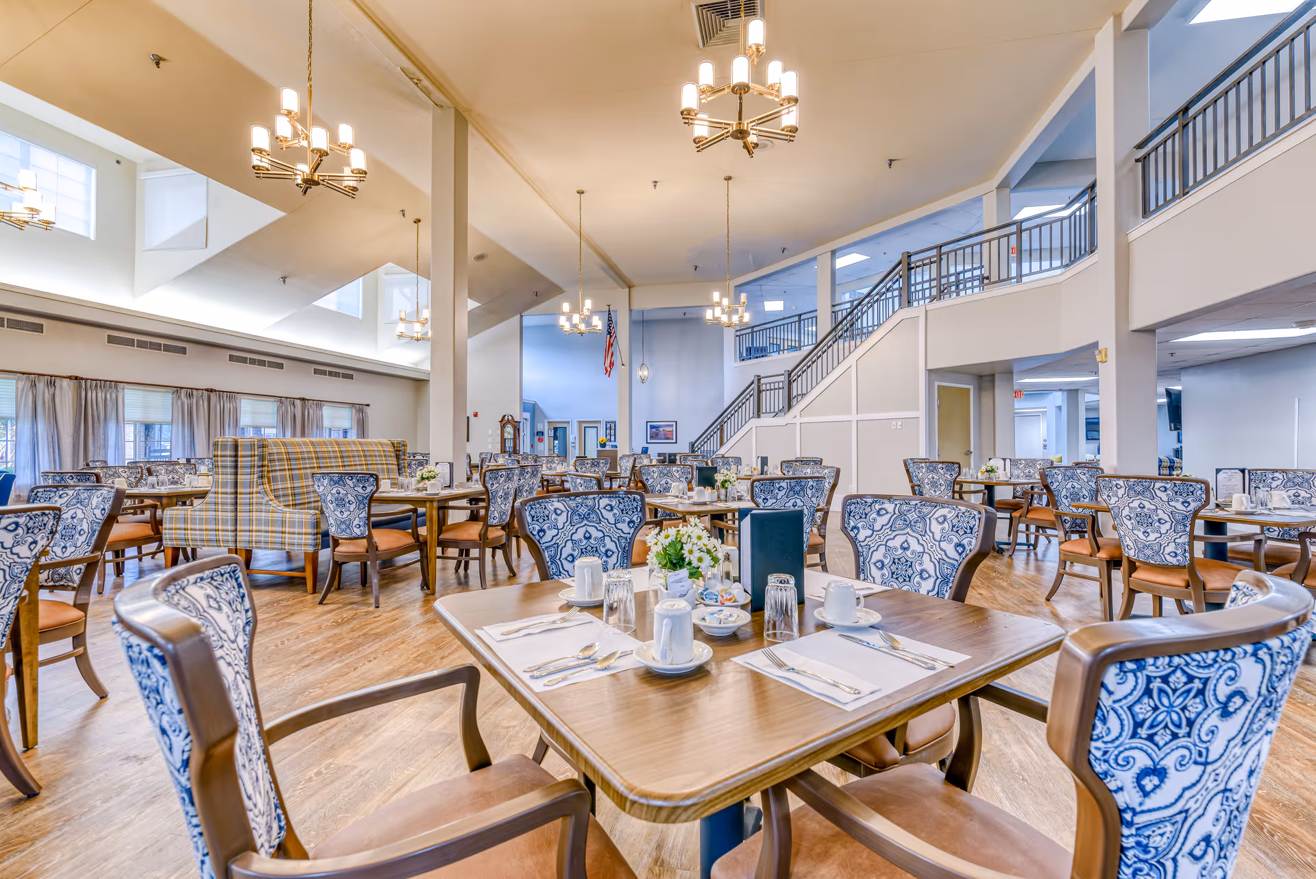 A spacious and well-lit dining room in a senior living facility with multiple tables and chairs arranged neatly. The chairs have blue and white patterned upholstery, and the tables are set with cups, glasses, utensils, and small flower arrangements. The room features high ceilings with hanging chandeliers, large windows with curtains, and a staircase leading to an upper level.
