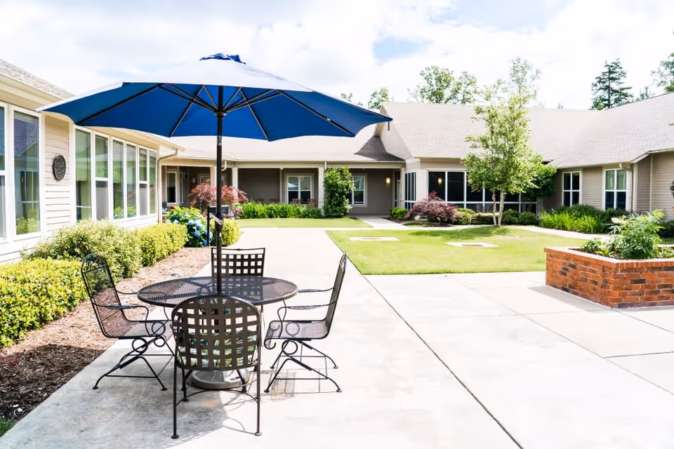 Outdoor courtyard area at Pinnacle Place Memory Care featuring a round metal table with four matching chairs and a large blue umbrella. The courtyard is surrounded by a single-story building with beige siding and multiple windows. There are green shrubs, small trees, and a brick planter box along the concrete walkway.