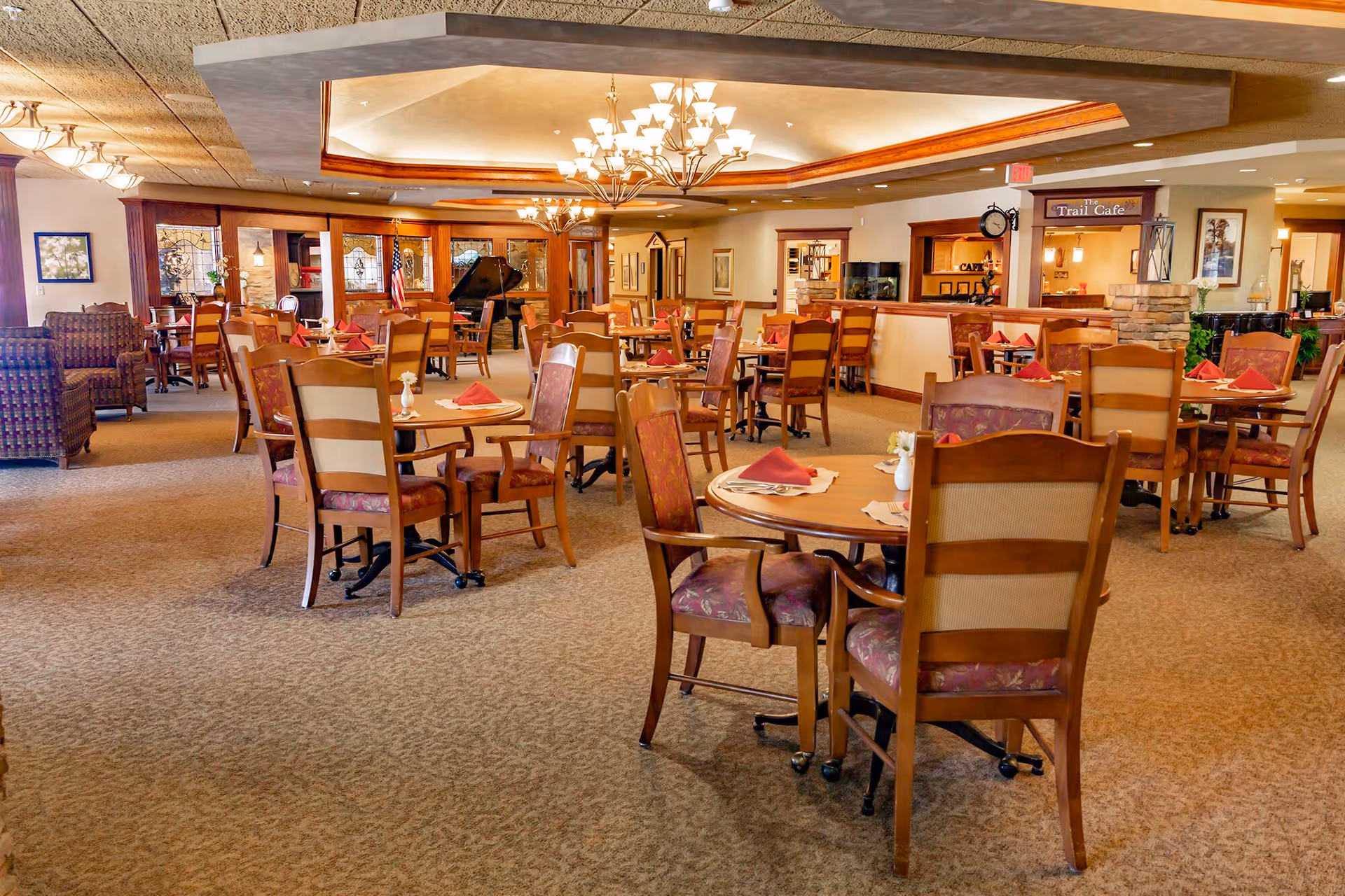 Spacious dining room with multiple round wooden tables and chairs, each table set with red napkins and small flower vases. The room features warm lighting with chandeliers, carpeted floor, and a cozy atmosphere. In the background, there is a grand piano, an American flag, and a sign that reads 'Trail Cafe'.