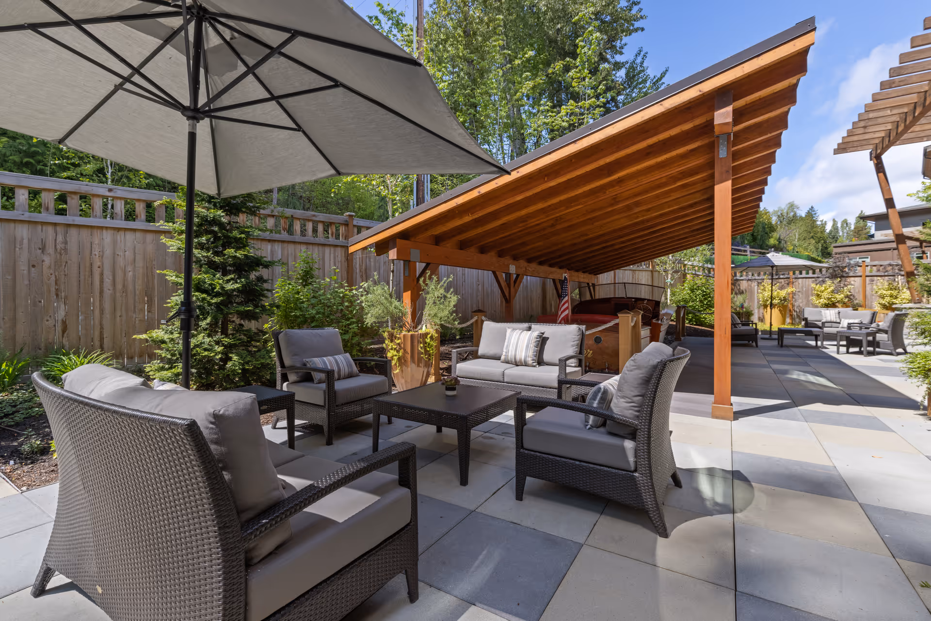 Outdoor patio area with cushioned wicker chairs and sofas arranged around a coffee table under a large umbrella and a wooden pergola. The space is surrounded by a wooden fence and greenery, with a clear blue sky overhead.