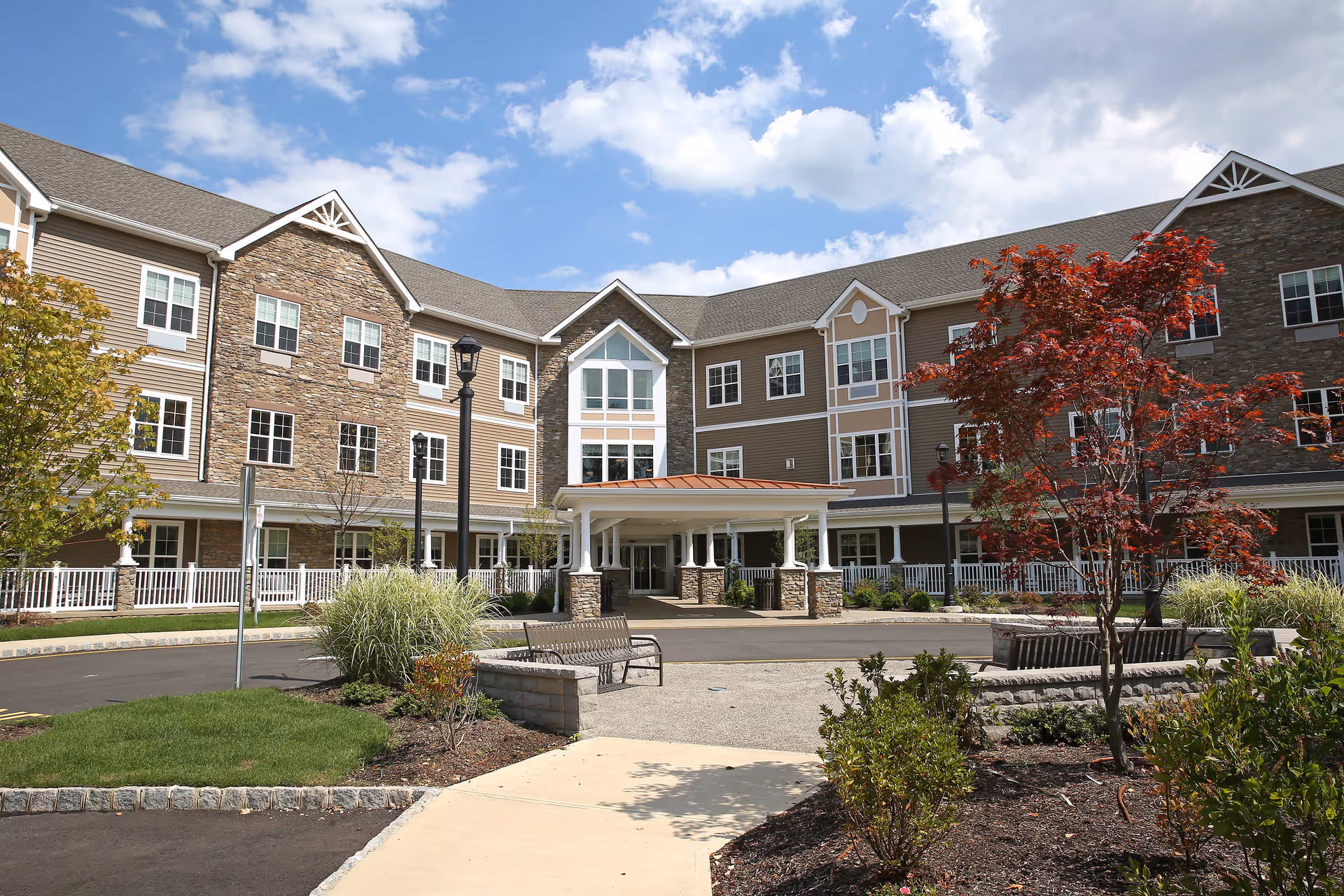 Front entrance of a three-story senior living building with stone and siding facade, covered drop-off, benches, and landscaped courtyard under a blue sky.