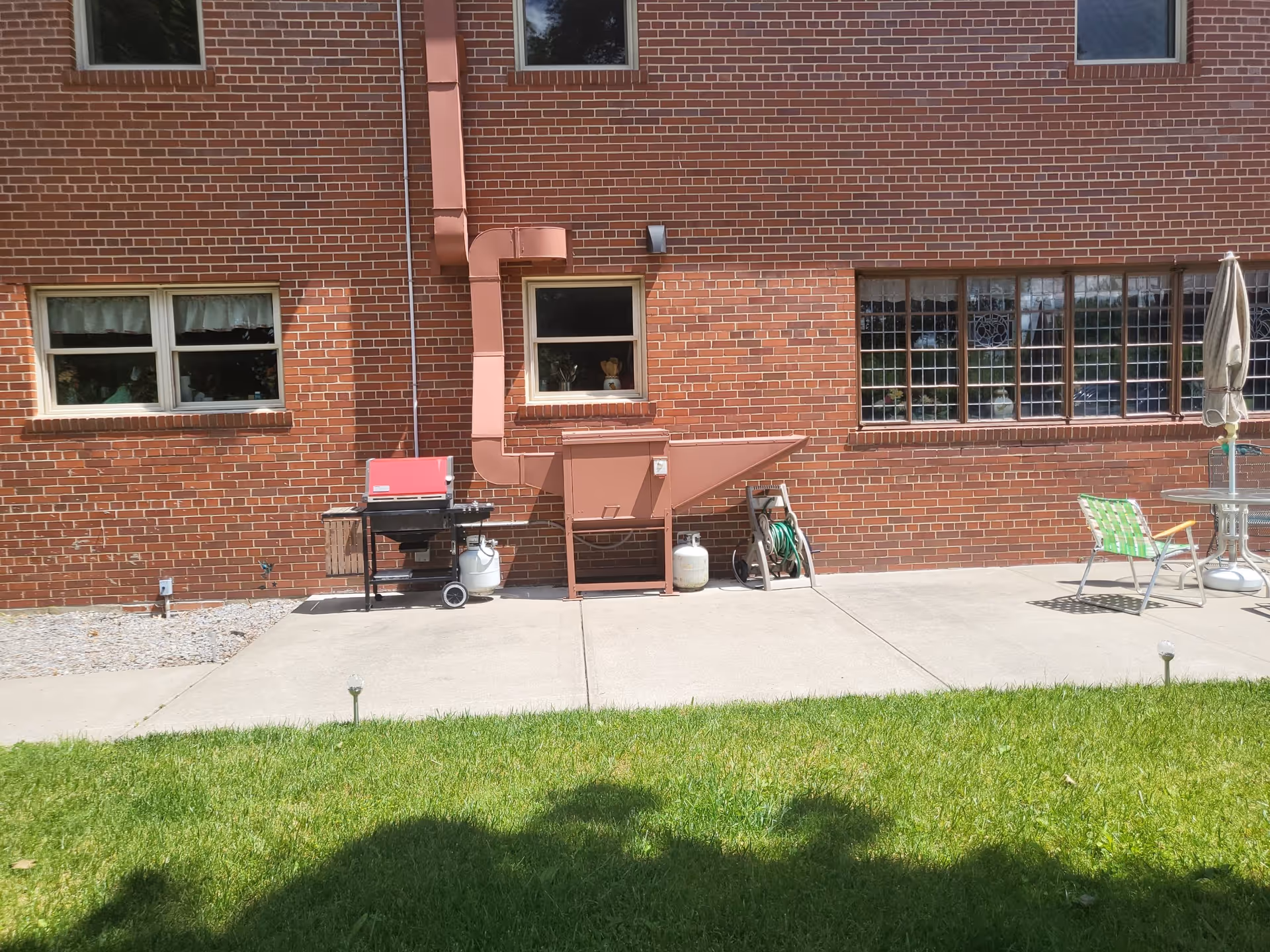 Outdoor patio area of a brick building with several windows. There is a red grill, a large brown metal vent or chute attached to the wall, a garden hose on a reel, a green and white lawn chair, a round glass table with a closed umbrella, and a concrete walkway with green grass in the foreground.