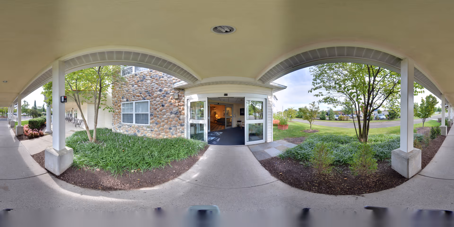 Covered walkway leading to the entrance of a building with stone and siding exterior walls, surrounded by landscaped greenery and trees under a cloudy sky.