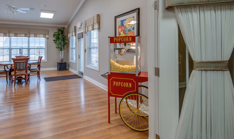 A bright room with wooden flooring featuring a red vintage popcorn machine filled with popcorn, a round wooden table with four chairs near large windows with blinds, a potted plant in the corner, and a framed colorful floral painting on the wall.