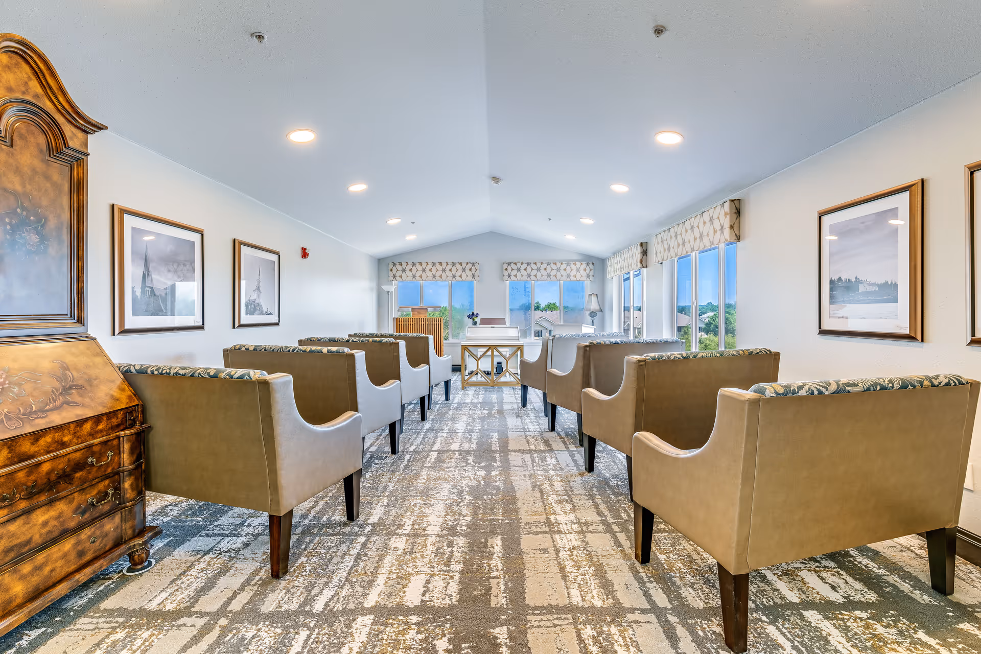 A bright, narrow room with rows of beige upholstered chairs facing a white piano and a small table at the far end. The room has large windows on the right side with patterned valances, framed pictures on the walls, and a wooden cabinet on the left. The ceiling is vaulted with recessed lighting.