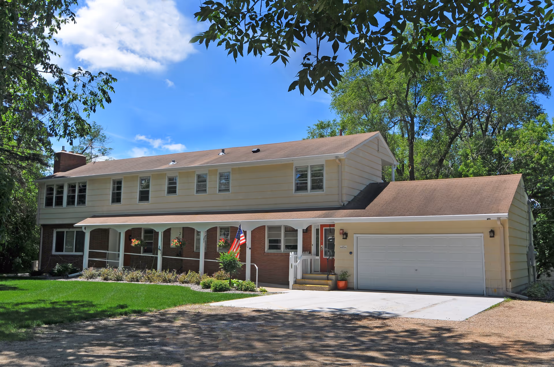 Exterior view of a two-story residential building with beige siding and a brown roof. The building has multiple windows, a covered porch with white pillars, hanging flower pots, an American flag, and a white garage door. The surrounding area includes green grass, trees, and a clear blue sky with some clouds.