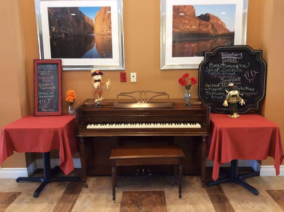 An upright piano with a matching bench is centered between two small tables covered with red tablecloths. Each table holds a small chalkboard menu and a decorative scarecrow figure. Above the piano are two framed landscape photographs of rocky cliffs reflected in water. The walls are painted a warm beige color.