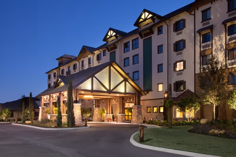 Exterior view of a multi-story senior living facility building at dusk with warm lights illuminating the entrance area featuring a covered driveway supported by stone pillars and landscaped greenery around.