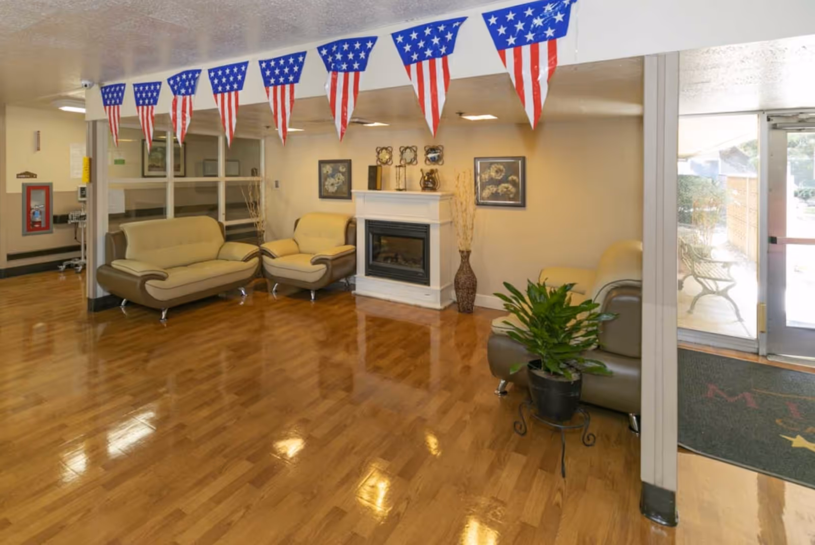 A clean and well-lit common area with polished wooden floors, beige and brown cushioned sofas arranged around a white fireplace, patriotic American flag bunting hanging from the ceiling, framed floral artwork on the walls, a potted plant on a small stand, and a glass door leading outside with a bench visible through the door.