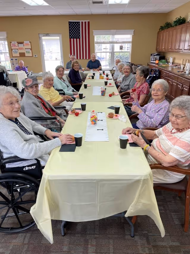A group of elderly people sitting around a long table covered with a yellow tablecloth in a well-lit room. They are smiling and appear to be enjoying a social gathering with snacks and drinks. An American flag hangs on the wall in the background, and there are windows letting in natural light. The room has wooden cabinets and a bulletin board on the wall.