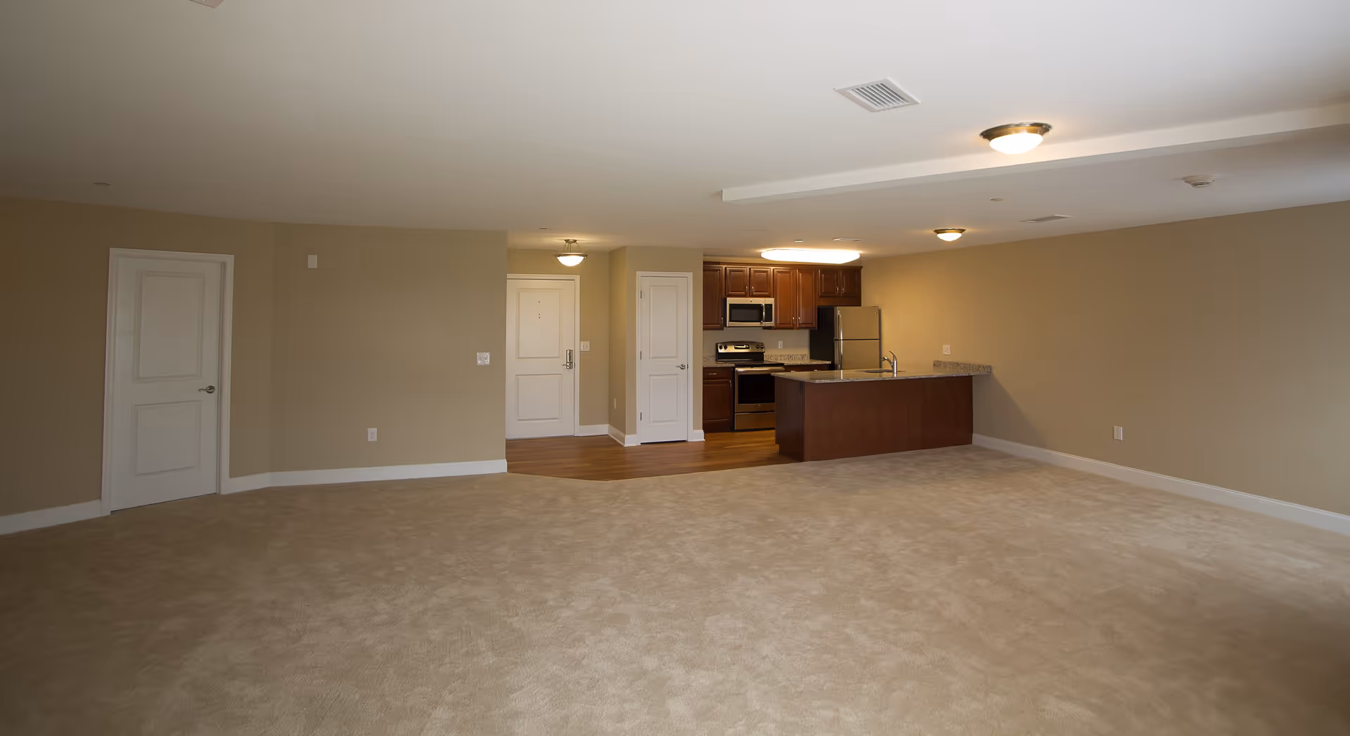 An empty apartment interior featuring a spacious living area with beige carpet flooring and beige walls. The kitchen area has wooden cabinets, stainless steel appliances including a refrigerator, stove, and microwave, and a granite countertop island with a sink. There are three white doors visible, one likely the entrance door and two others possibly for closets or rooms. The ceiling has multiple light fixtures and air vents.