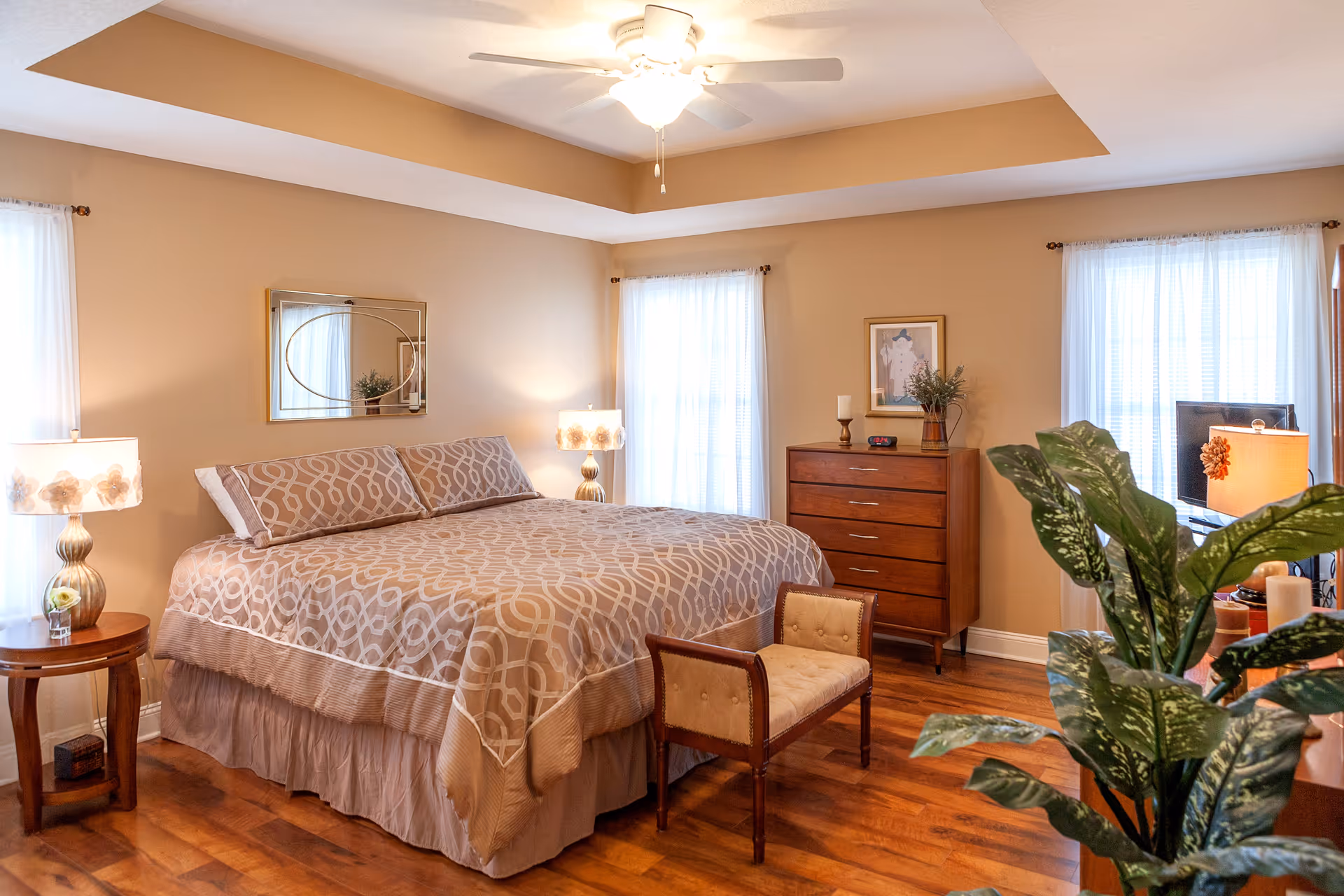A well-lit bedroom with a large bed covered in a beige patterned bedspread, two bedside tables with lamps, a wooden dresser with decorative items, a small upholstered bench at the foot of the bed, hardwood floors, and large windows with sheer white curtains. A green leafy plant is visible in the foreground.