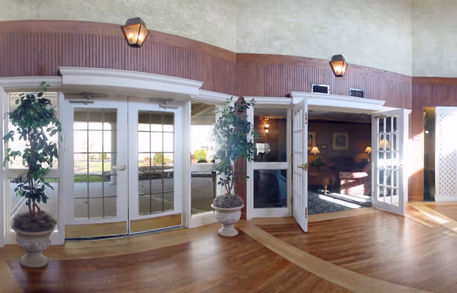 Entrance area with glass double doors and adjacent open French doors leading into a cozy living room with lamps and furniture. Two potted plants flank the doors, and the floor is wood with a light-colored wall and wooden paneling above the doors.