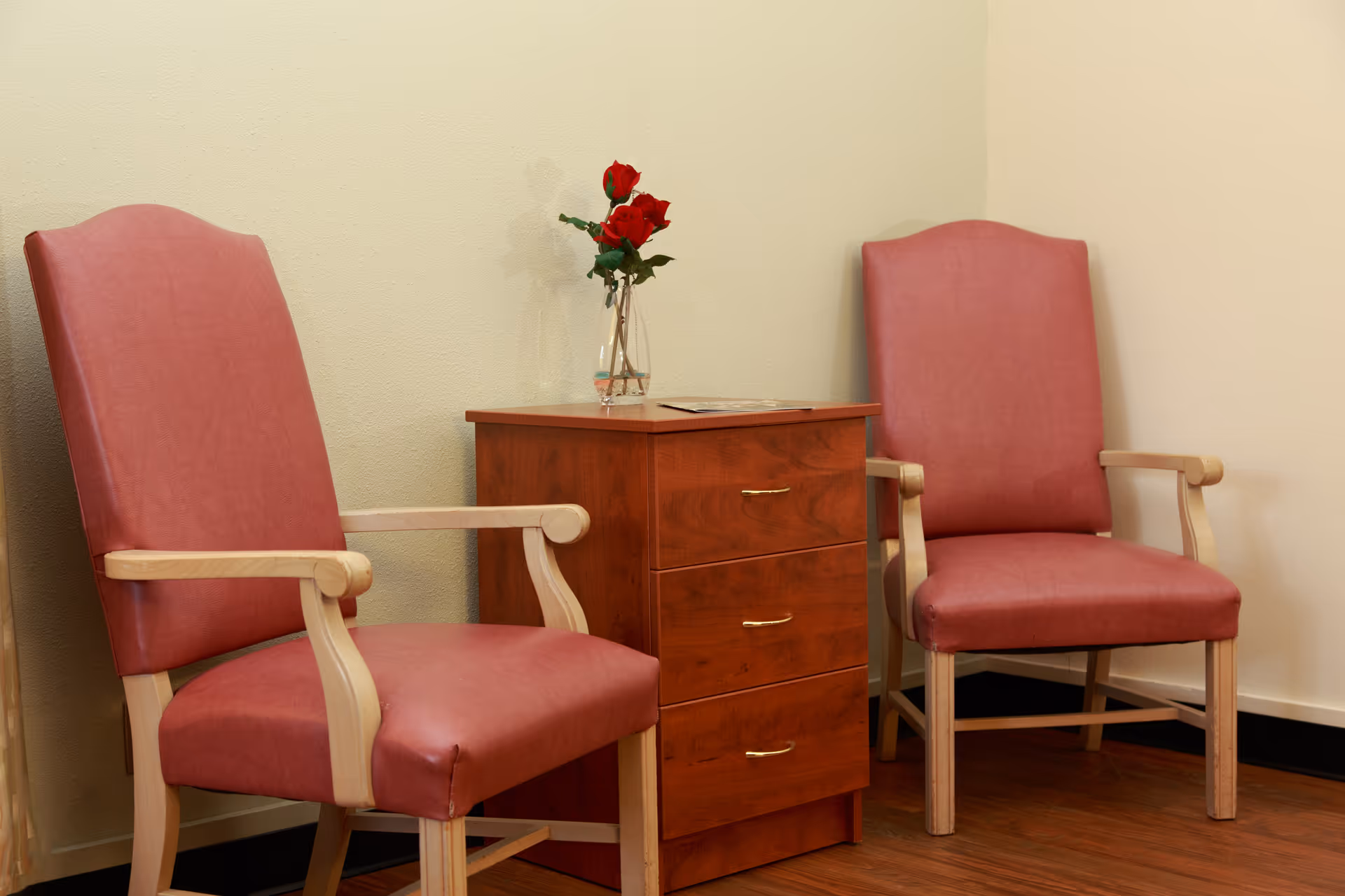 Two red cushioned armchairs with wooden frames are positioned on either side of a small wooden cabinet with three drawers. A clear vase with red roses is placed on top of the cabinet against a plain beige wall.