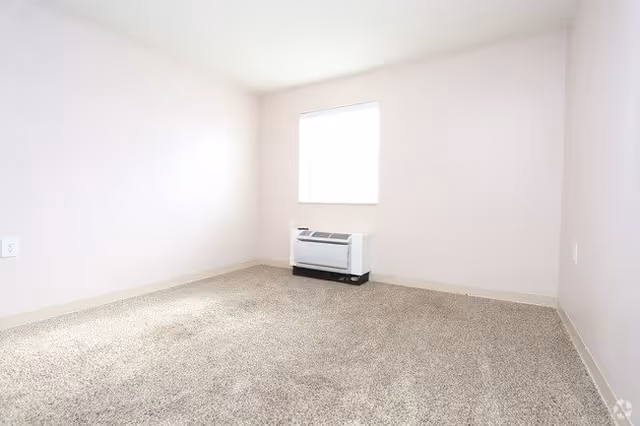 Empty room with beige carpet, light pink walls, a single window with blinds, and a white heating or air conditioning unit beneath the window.