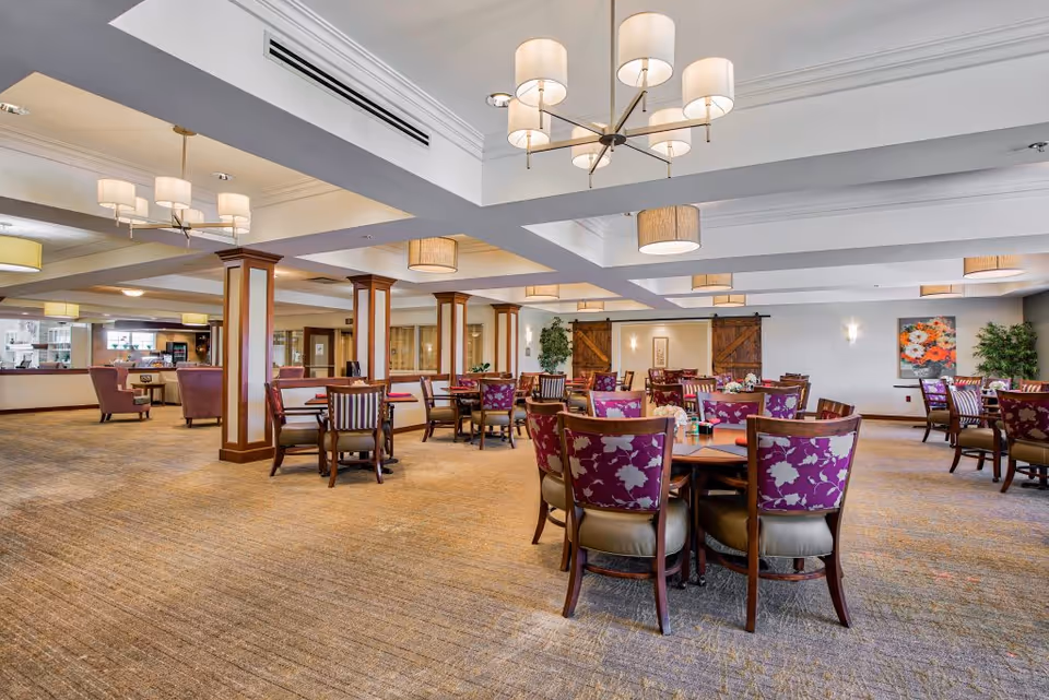 A spacious dining room with multiple round tables surrounded by chairs with purple floral upholstery. The room features beige carpeting, wooden columns, and modern ceiling light fixtures. There are decorative plants and artwork on the walls, and a counter area is visible in the background.