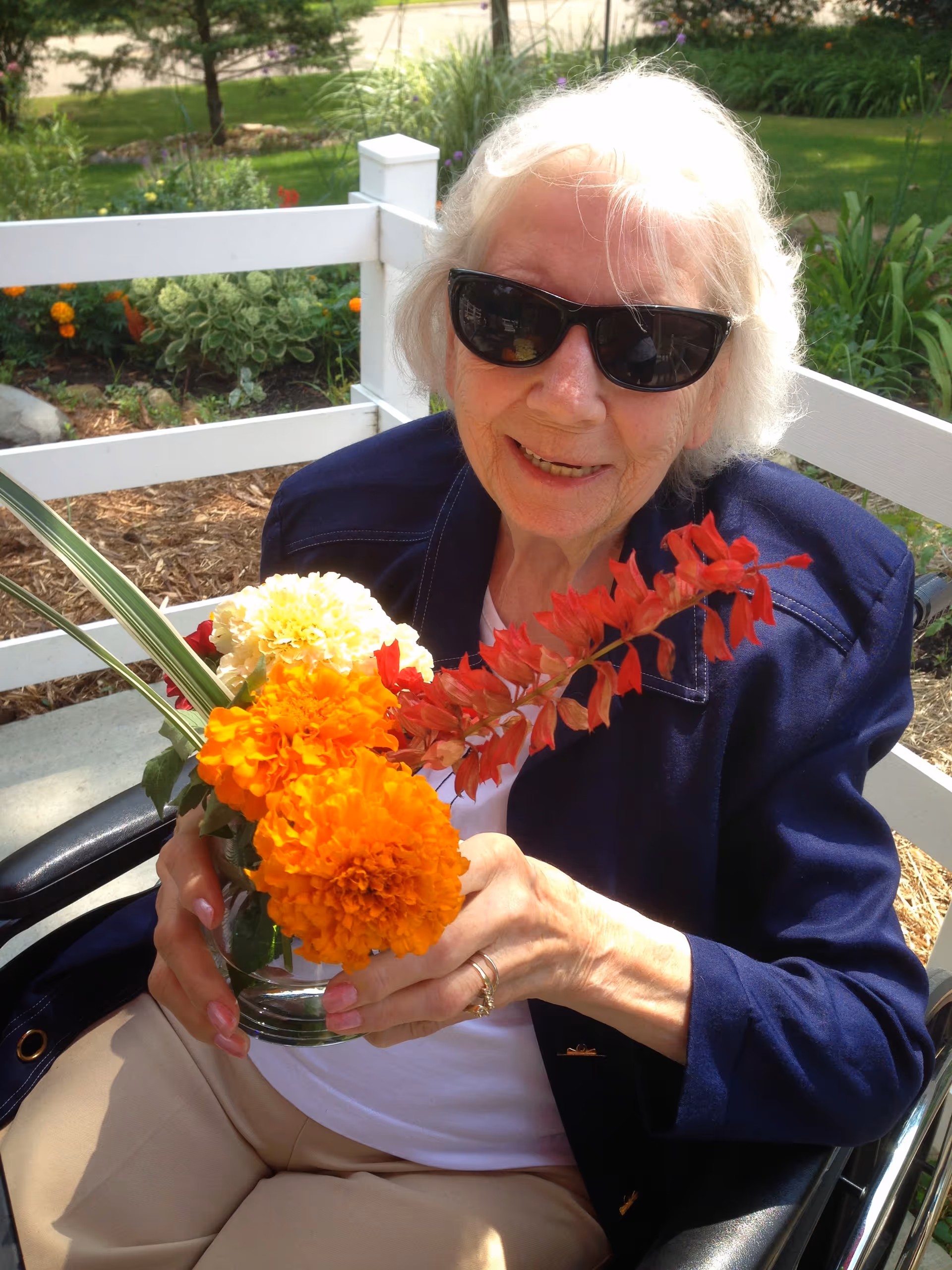 An elderly woman wearing sunglasses and a navy blue jacket is sitting outdoors in a wheelchair, holding a small glass vase with bright orange, yellow, and red flowers. Behind her is a white wooden fence and a garden with green plants and flowers.