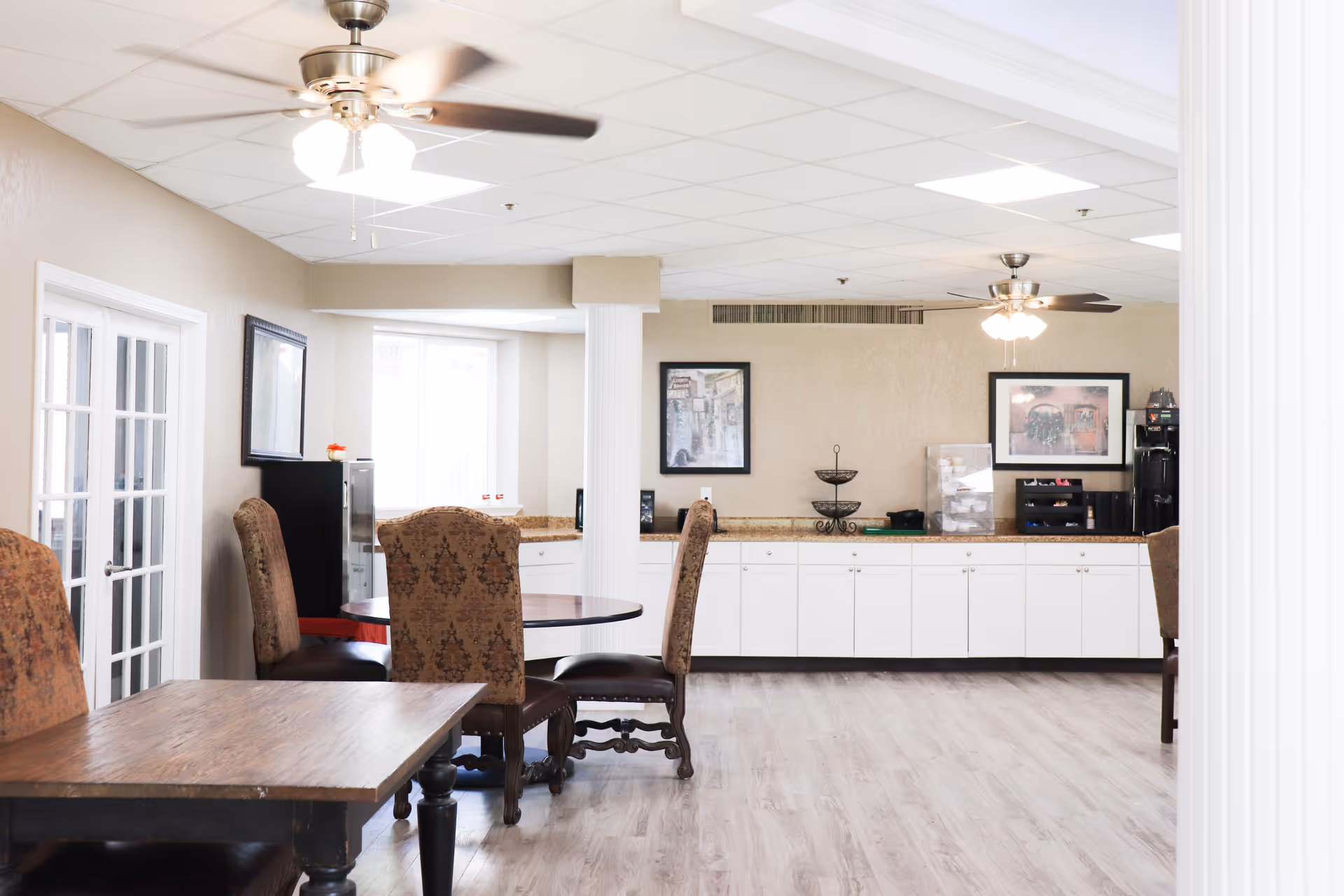 Bright communal dining area with wooden tables, upholstered chairs, white cabinets along the back wall and ceiling fans.