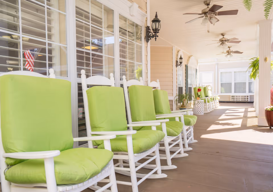 A bright and spacious covered porch with white rocking chairs featuring green cushions lined up along the side of a building. The porch has ceiling fans, hanging plants, and decorative wall lanterns, creating a welcoming outdoor seating area.