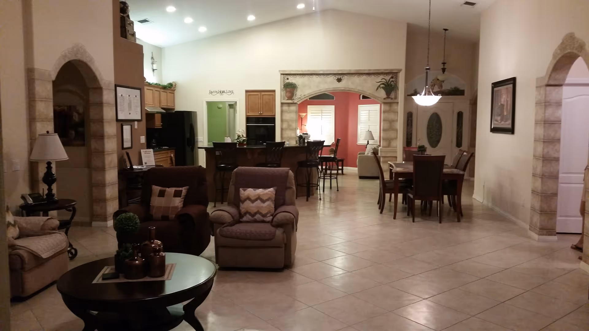Interior view of a senior living facility showing a spacious open area with a living room, dining area, and kitchen. The living room has comfortable armchairs and a round coffee table with decorative items. The dining area has a wooden table with chairs and a hanging light fixture above. The kitchen features wooden cabinets, a black refrigerator, and a counter with bar stools. The walls are painted in neutral tones with some red accent walls and decorative arches.