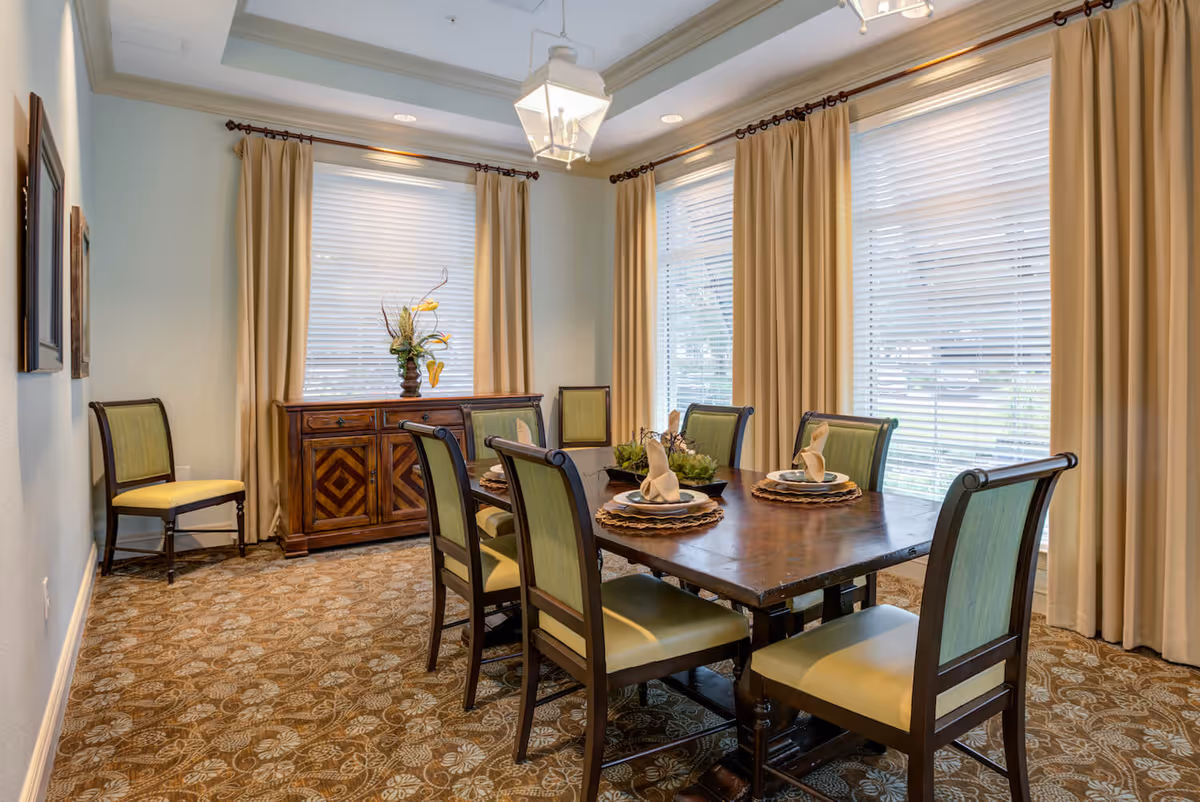A dining room with a wooden table set for four with plates, napkins, and decorative centerpieces. The room has six green cushioned chairs around the table and one additional chair against the wall. There are large windows with beige curtains and blinds, a patterned carpet, and a wooden sideboard with a vase of flowers.