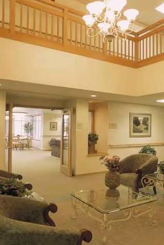 A spacious senior living lobby with seating, glass coffee table, plants, chandelier, artwork, and upper balcony.