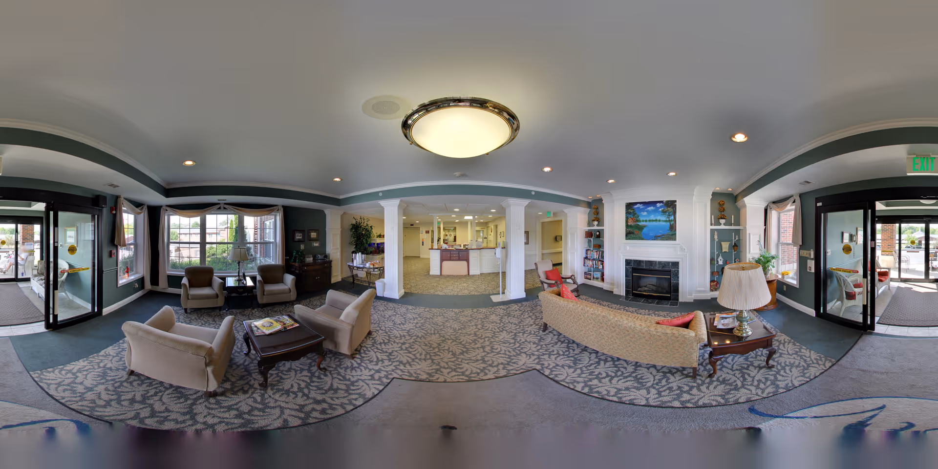 A spacious and well-lit retirement community common area with comfortable seating including armchairs and sofas arranged around coffee tables. The room features large windows, a fireplace with a painting above it, built-in shelves, and a reception desk in the background. The decor includes plants, lamps, and patterned carpeting.