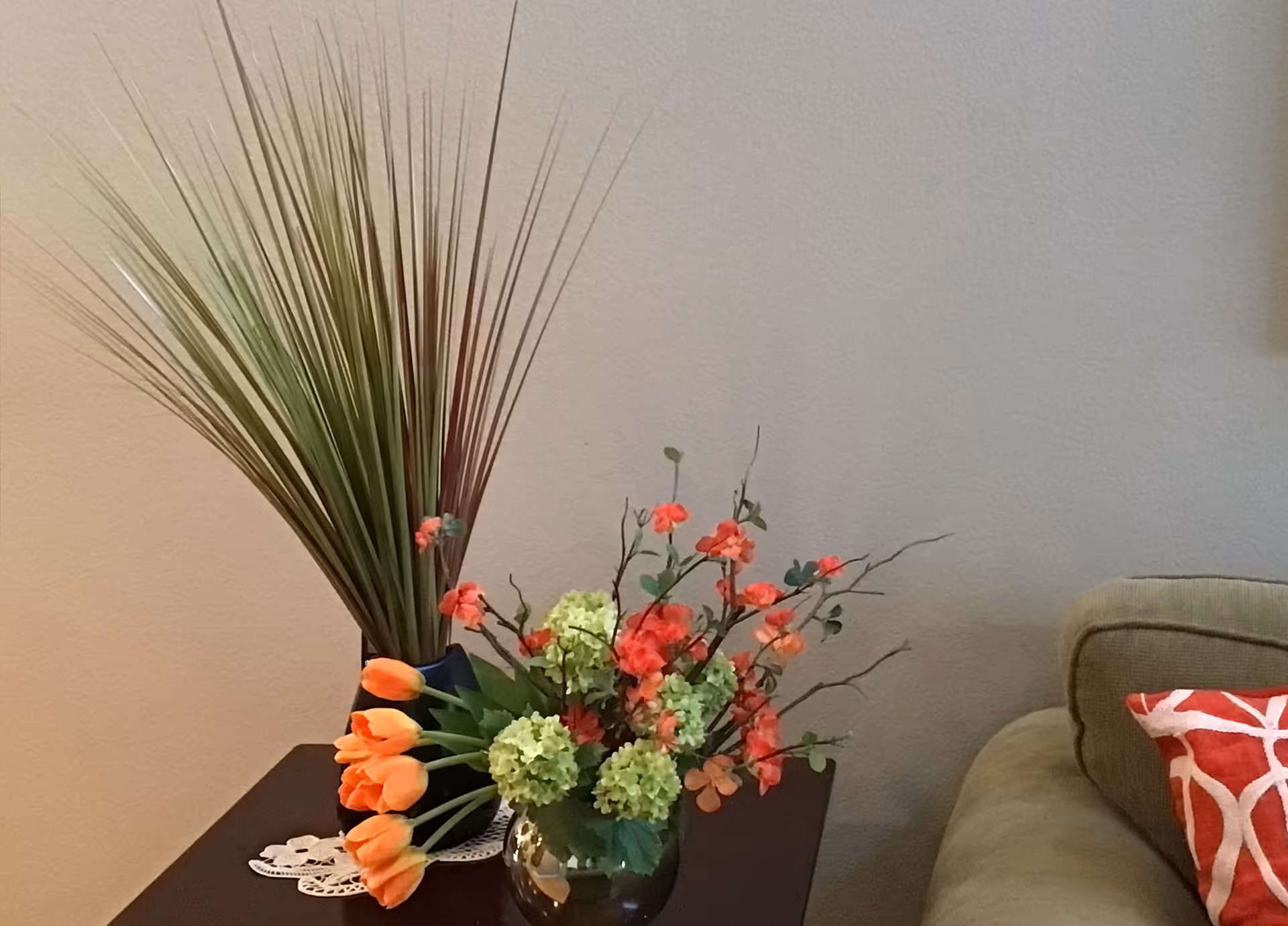 A close-up view of a dark wooden side table next to a beige sofa with a red and white patterned pillow. On the table, there are two floral arrangements: one with tall green and brown grass-like plants in a black vase, and another with orange tulips and green and orange flowers in a round glass vase. The background is a plain beige wall.