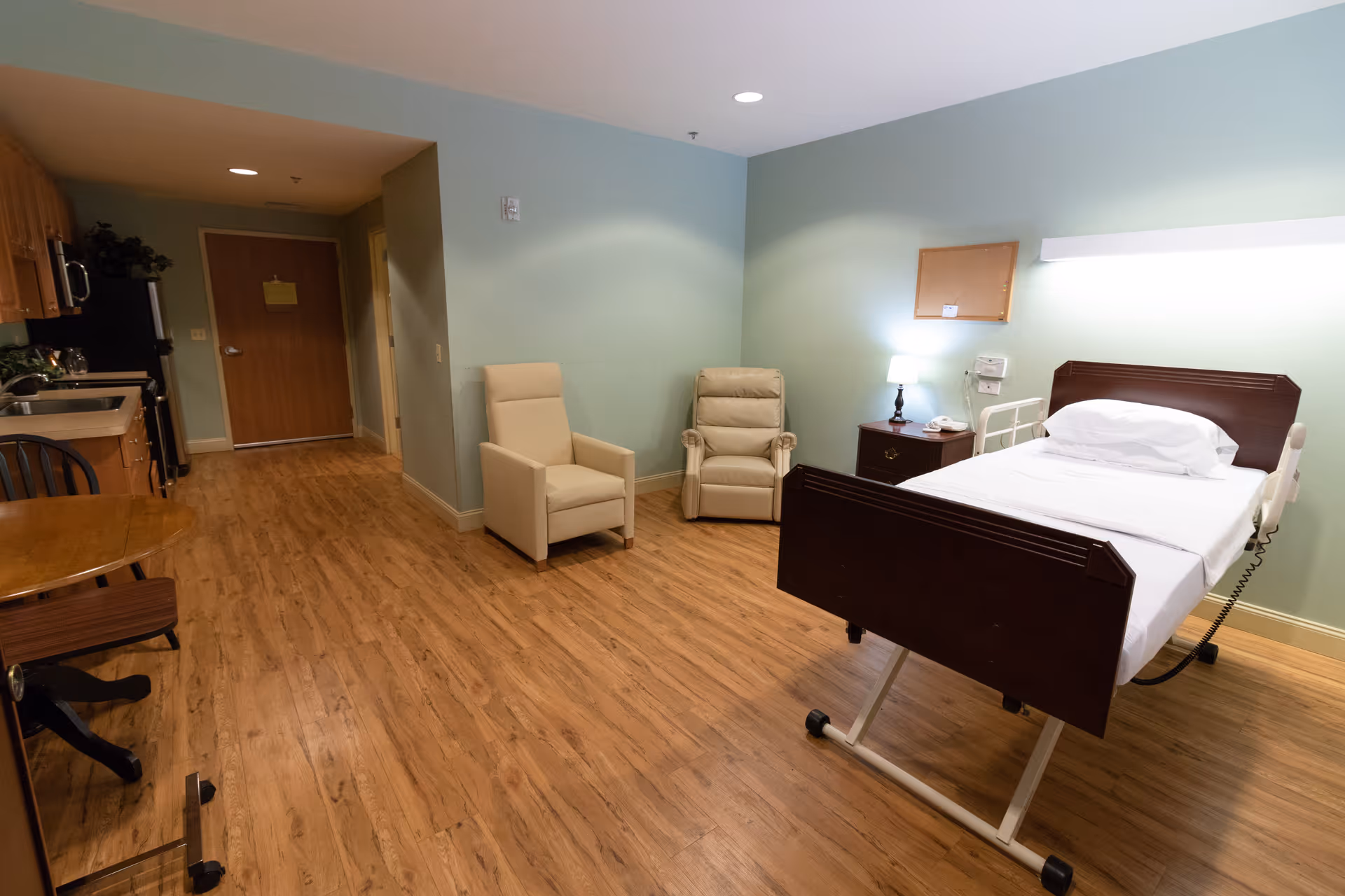 A spacious room in a care facility with a single hospital bed on the right side, two beige armchairs against a light green wall, a small wooden nightstand with a lamp and a telephone, and a kitchenette area with wooden cabinets, a sink, and a refrigerator on the left side. The floor is wooden, and the room has a calm and clean atmosphere.