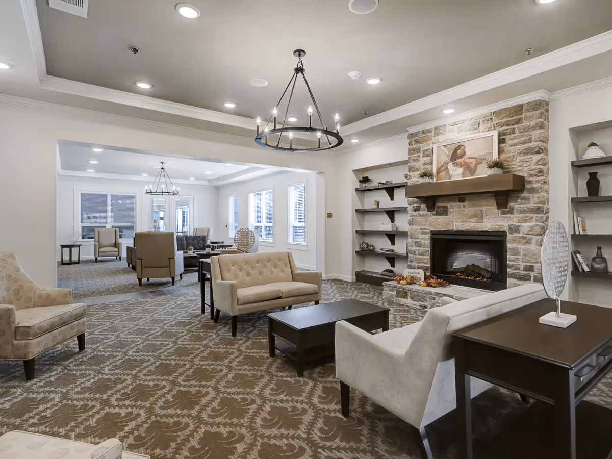 A spacious and well-lit living room area with beige upholstered chairs and sofas arranged around dark wood coffee and side tables. The room features a stone fireplace with a mantle decorated with plants and a framed picture above it. Built-in shelves flank the fireplace, holding decorative items and books. The ceiling has recessed lighting and a modern chandelier. Large windows allow natural light to fill the space.