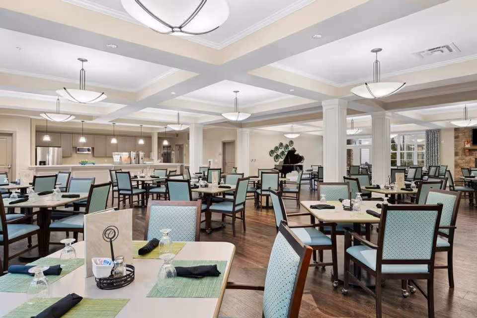 Bright, spacious dining room with multiple set tables, teal-upholstered chairs, and a serving counter and piano in the background.