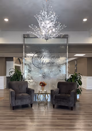 Lobby lounge with two gray armchairs flanking a small table in front of a glass water wall engraved 'Hampton Manor' and a decorative chandelier overhead.