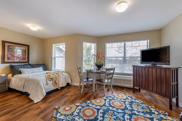 A bright and spacious senior living bedroom with a large bed on the left side, a small round dining table with four chairs near the windows, a wooden cabinet with a flat-screen TV on the right, and a colorful patterned rug on the wooden floor.