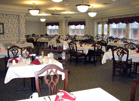 Spacious dining room with multiple tables set with white tablecloths, red napkins, wooden chairs, and ceiling lights.