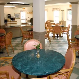 Interior view of a dining area with multiple round tables and chairs. The tables have greenish marble-like surfaces and some have small decorative plants. The room has large windows with floral curtains allowing natural light in. The floor is checkered with green and white tiles, and there is a counter area in the background.