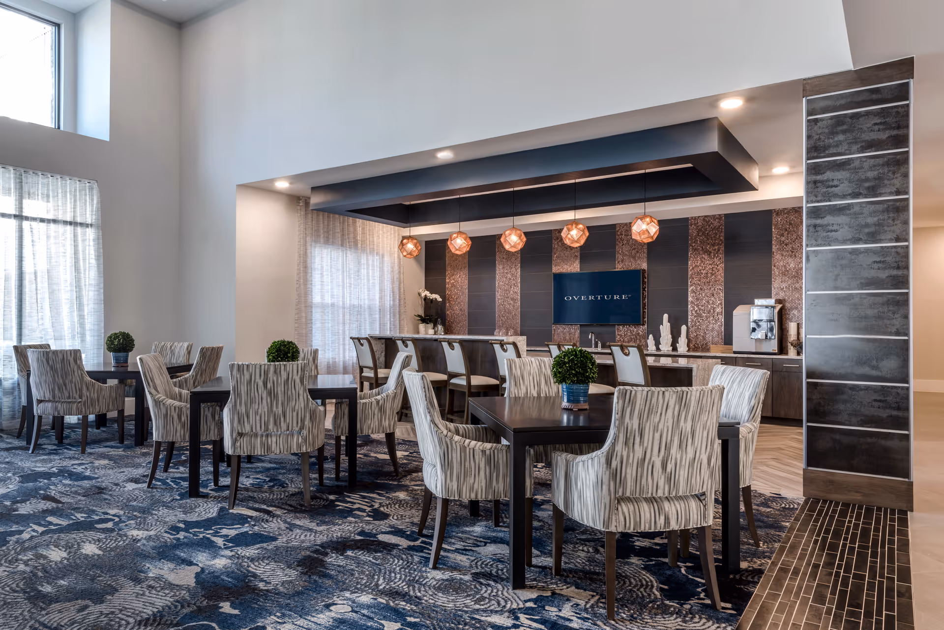 A modern dining area with multiple tables and upholstered chairs arranged on a patterned blue carpet. The back wall features a bar counter with high chairs, pendant lights hanging from the ceiling, and a TV screen displaying the word 'OVERTURE'. There are decorative plants on the tables and a coffee machine on the counter to the right.