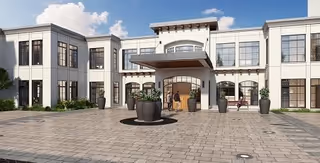 Front exterior view of a two-story senior living facility building with large windows, a covered entrance, and several large planters on a paved driveway under a partly cloudy sky.