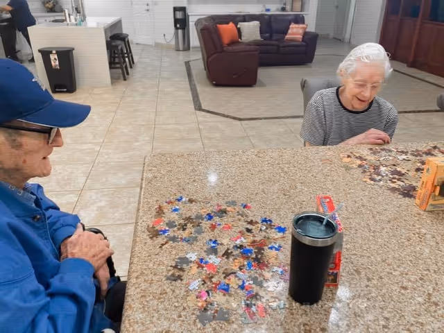 Two elderly individuals sitting at a large table working on separate jigsaw puzzles in a spacious, tiled common area with a brown leather couch and colorful pillows in the background.