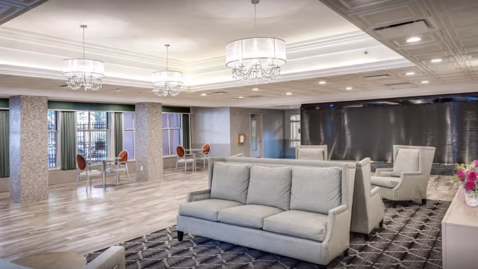 A spacious and well-lit common area with modern decor, featuring light gray sofas and armchairs arranged on a patterned rug. The room has large windows with green curtains, multiple chandeliers hanging from a coffered ceiling, and small tables with red chairs near the windows.