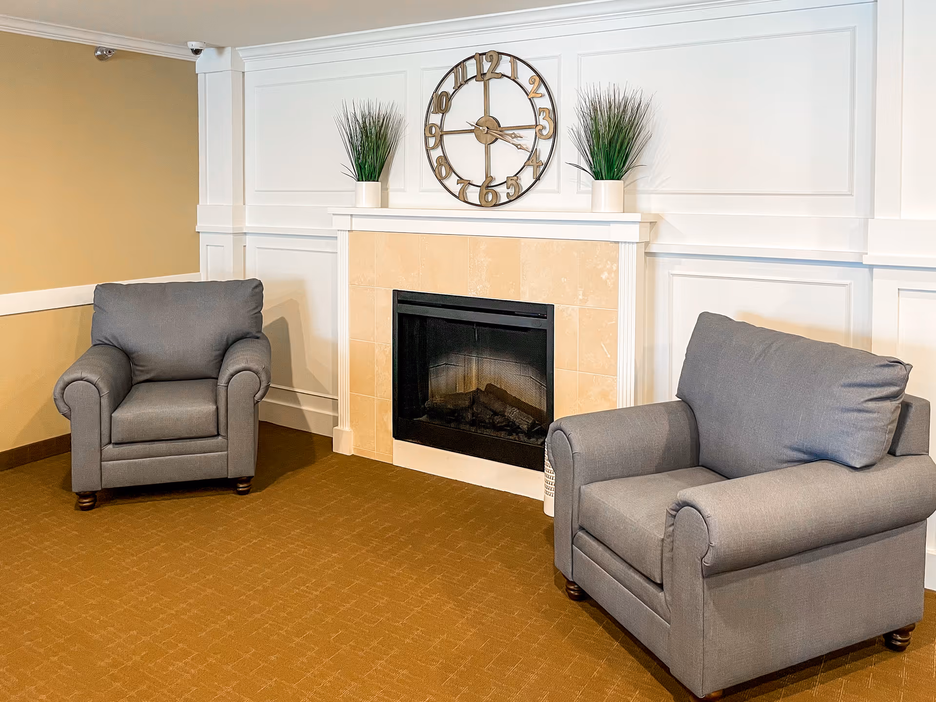 A cozy seating area with two gray armchairs facing a beige tiled fireplace. Above the fireplace is a large decorative clock and two potted plants on the mantel. The walls are white with panel molding, and the floor is carpeted in a brown color.