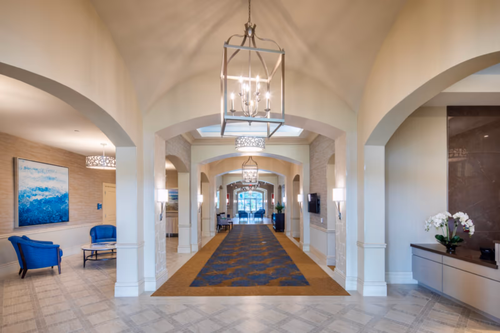 A spacious and well-lit hallway in a senior living facility with arched doorways, a patterned carpet runner, and modern chandeliers hanging from a vaulted ceiling. To the left, there is a seating area with blue chairs and a coffee table beneath a large blue and white abstract painting. On the right, there is a countertop with a vase of white orchids and a dark marble backsplash.