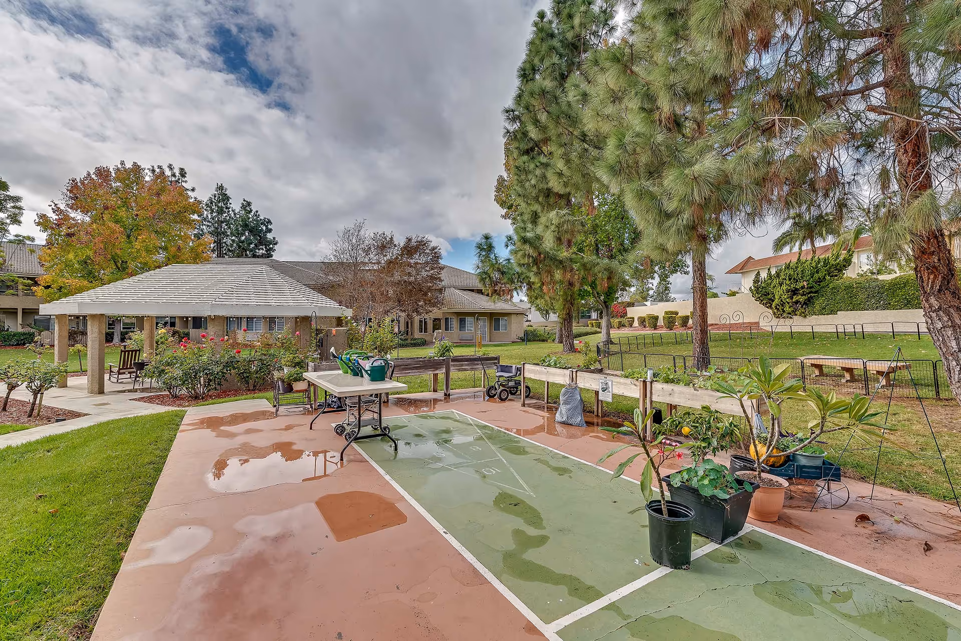 Outdoor garden area at Arcadia Place Senior Living featuring a shuffleboard court with potted plants, a table with watering cans, a gazebo with chairs, and surrounding trees and greenery under a cloudy sky.