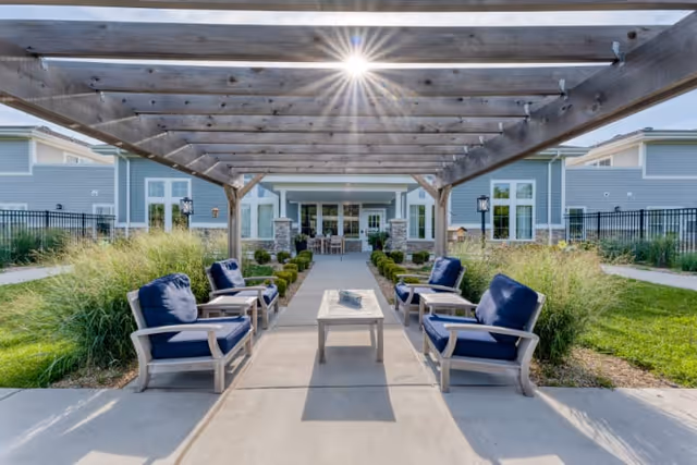 Outdoor seating area with four cushioned chairs arranged around a rectangular table under a wooden pergola. The area is surrounded by green grass and ornamental grasses, with a building featuring large windows and a patio in the background. The sun is shining brightly above the pergola.