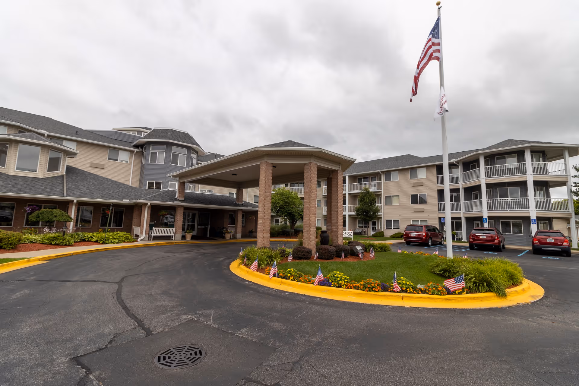 Exterior view of Independence Village of Saginaw showing the entrance with a covered drop-off area supported by brick columns, a circular driveway with a landscaped island featuring flowers and small American flags, and a tall flagpole with an American flag. The building is multi-story with beige and gray siding, balconies, and several parked cars in front.