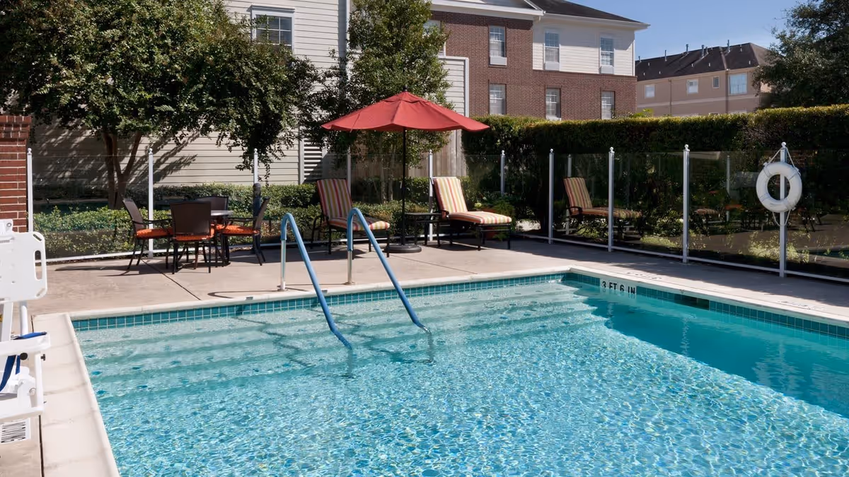 Outdoor swimming pool with clear blue water, metal handrails, and a safety ring on a glass fence. Surrounding the pool are lounge chairs with striped cushions, a red umbrella, a table with chairs, and greenery including trees and bushes. Residential buildings are visible in the background.