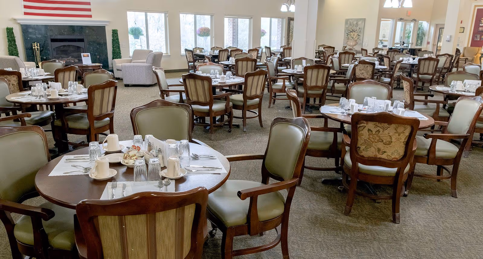 A spacious dining room with multiple round wooden tables set with cups, glasses, plates, and silverware. The chairs around the tables have wooden frames with green and floral upholstery. Large windows let in natural light, and there is a fireplace with an American flag above it in the background.