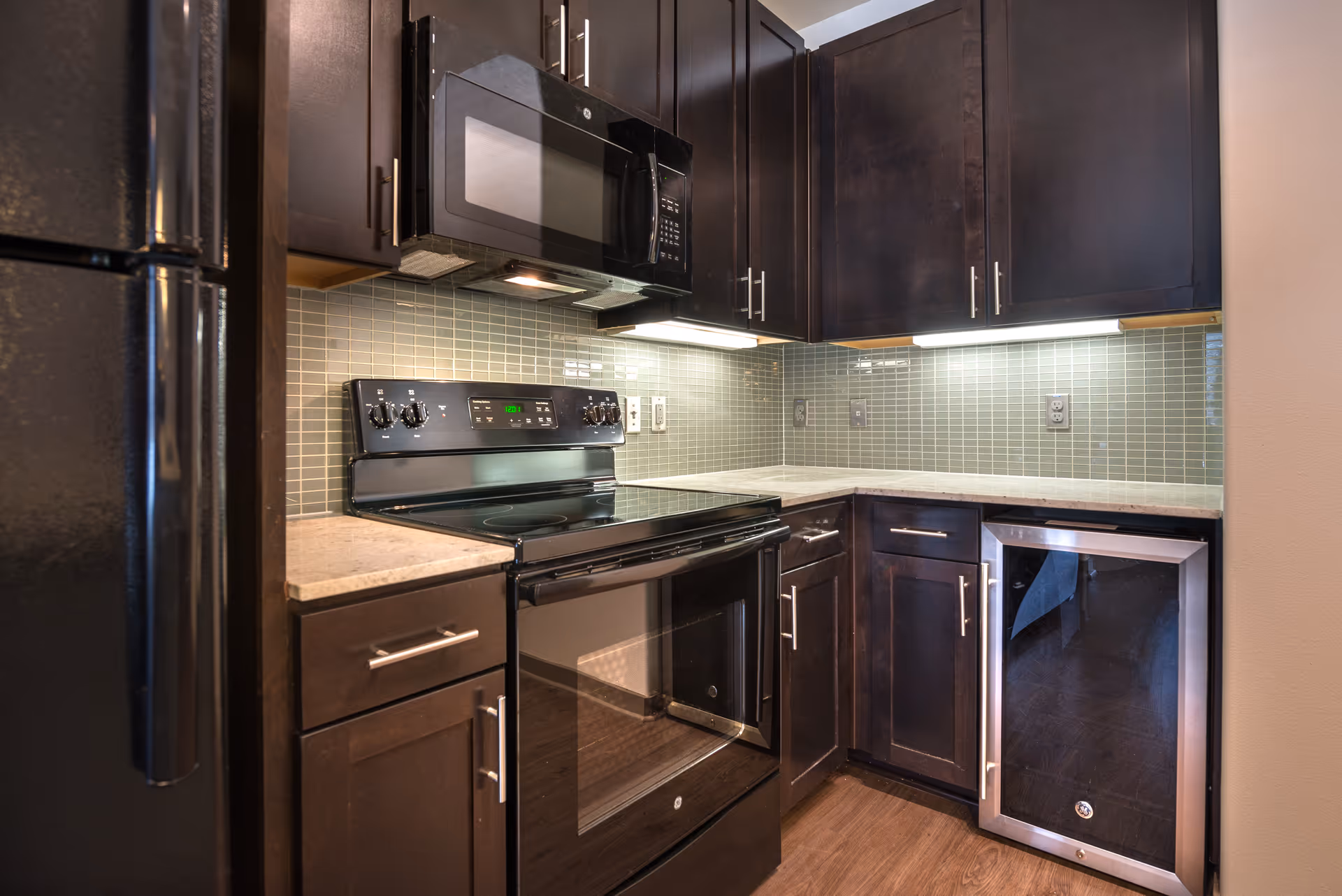 Modern kitchen with dark wood cabinets, a black electric stove with oven, a black microwave above the stove, a black refrigerator on the left, and a small wine cooler or beverage fridge on the right under the counter. The backsplash is made of small rectangular light gray tiles, and the countertops are light-colored.