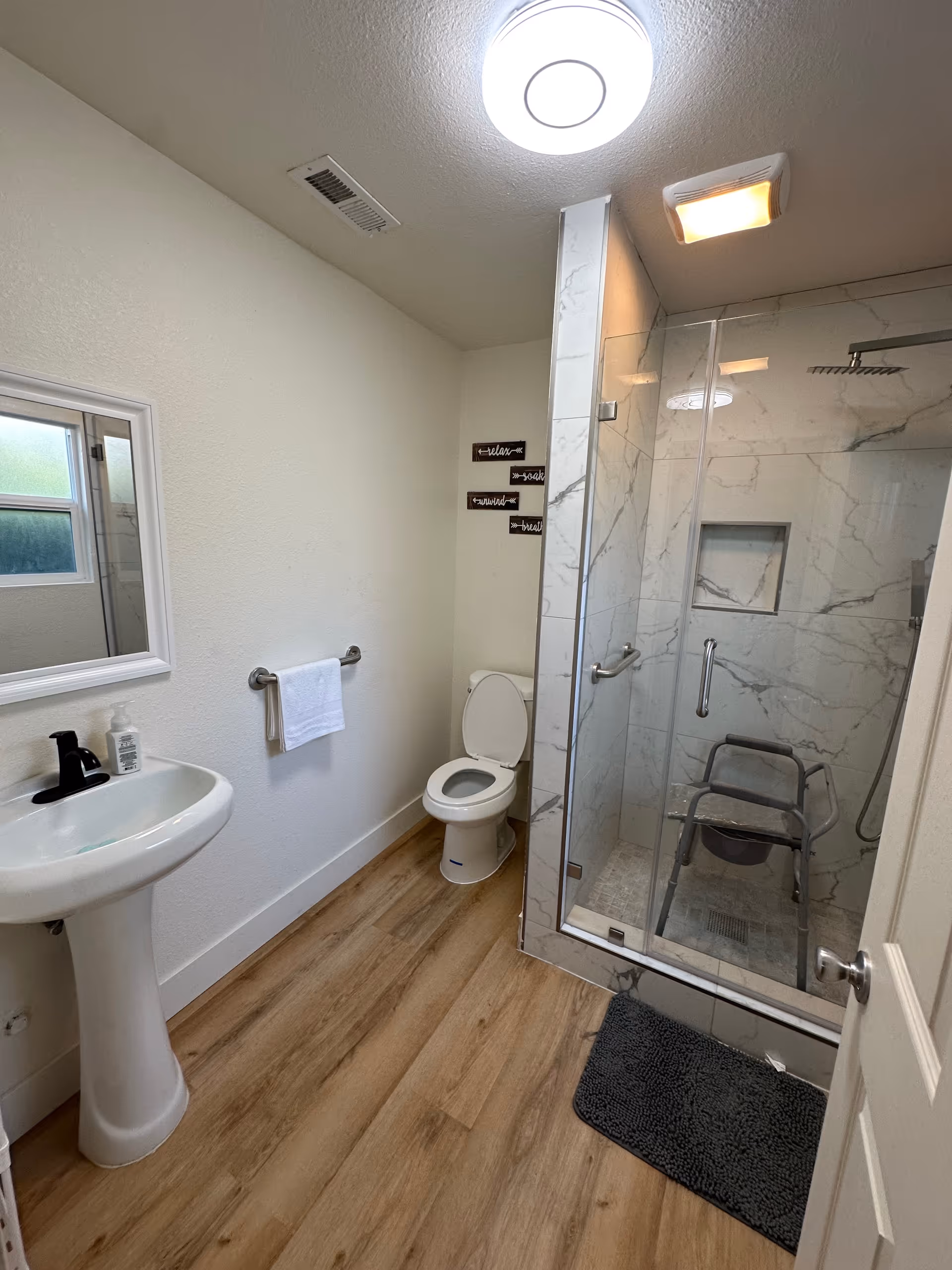 A clean bathroom with a white pedestal sink, a toilet with the lid open, and a glass-enclosed shower with marble-patterned walls. Inside the shower is a gray shower chair. The floor is wood-style laminate, and there is a towel hanging on a bar next to the sink. The ceiling has a round light fixture and a vent.